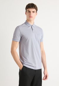 Polo de color gris claro con cuello clásico, mangas cortas y botonera de tres botones; presenta un sutil logotipo en el pecho.