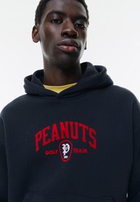 Hanorac bleumarin cu grafică "PEANUTS GOLF TEAM" în roșu, cu un logo rotund și un buzunar tip cangur. Fabrică moale și glugă ajustabilă.