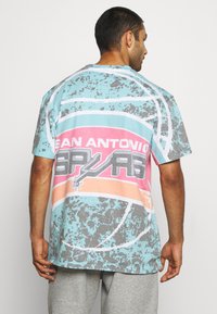 Mand iført en flerfarvet San Antonio Spurs T-shirt med et basketballgrafik, vendt væk mod en ensfarvet baggrund.