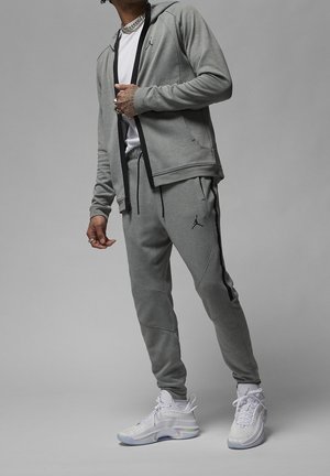 Persoon draagt een grijze zip-up hoodie en joggingbroek met zwarte biezen en logo's, witte sneakers en gelaagde zilveren kettingen om de hals.