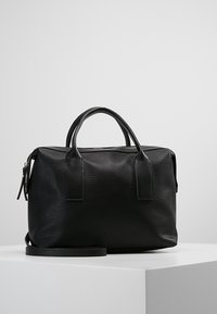 Svart strukturerad läderhandväska med dubbla handtag och en axelrem, uppställd på en vit pelare mot en slät ljusgrå bakgrund.