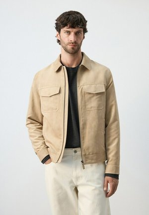 Hombre con chaqueta beige con cremallera sobre camisa negra y pantalones blancos, de pie contra un fondo claro y liso.