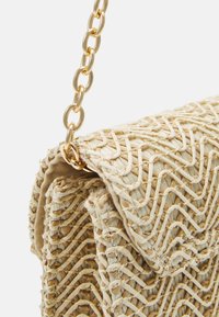 Sac à main beige tissé avec motif chevron et bandoulière en chaîne dorée en gros plan sur un fond de tissu texturé.