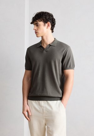 Fiatal férfi göndör sötét hajjal, olívazöld rövid ujjú pólóinget és krémszínű nadrágot viselve, egyik kezét zsebében tartva áll.