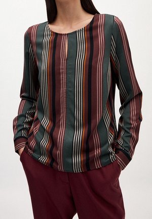 Pitkähihainen pusero, jossa on pystysuoria raitoja vihreässä, burgundinpunaisessa, mustassa, beigen ja oranssin sävyissä, yhdistettynä burgundinpunaisiin housuihin.