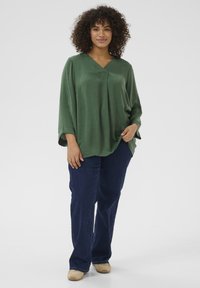 Blusa verde, holgada, con escote en V y mangas de tres cuartos, combinada con jeans rectos de color azul oscuro y zapatos beige.