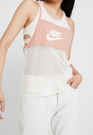 Osoba nosząca biały i różowy siateczkowy top Nike na białym sportowym biustonoszu, w połączeniu z białymi spodniami, z jedną ręką w kieszeni.