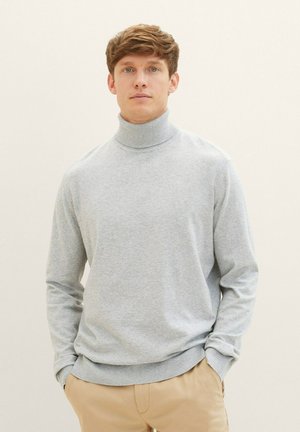 Un pull col roulé gris clair en tissu doux, doté de manches longues et de bords côtes aux poignets et à l'ourlet, porté avec un pantalon beige.