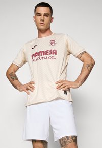 Vyras, dėvintis smėlio spalvos Villarreal CF futbolo marškinėlius ir baltus šortus, stovi pasitikėdamas savimi, laikydamas rankas ant klubų prieš vienodą foną.