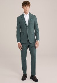 Zöldeskék öltöny, texturált felülettel, testreszabott fazonnal, kivágott hajtókákkal és két elülső zsebbel, fehér inggel és sötét cipővel párosítva.