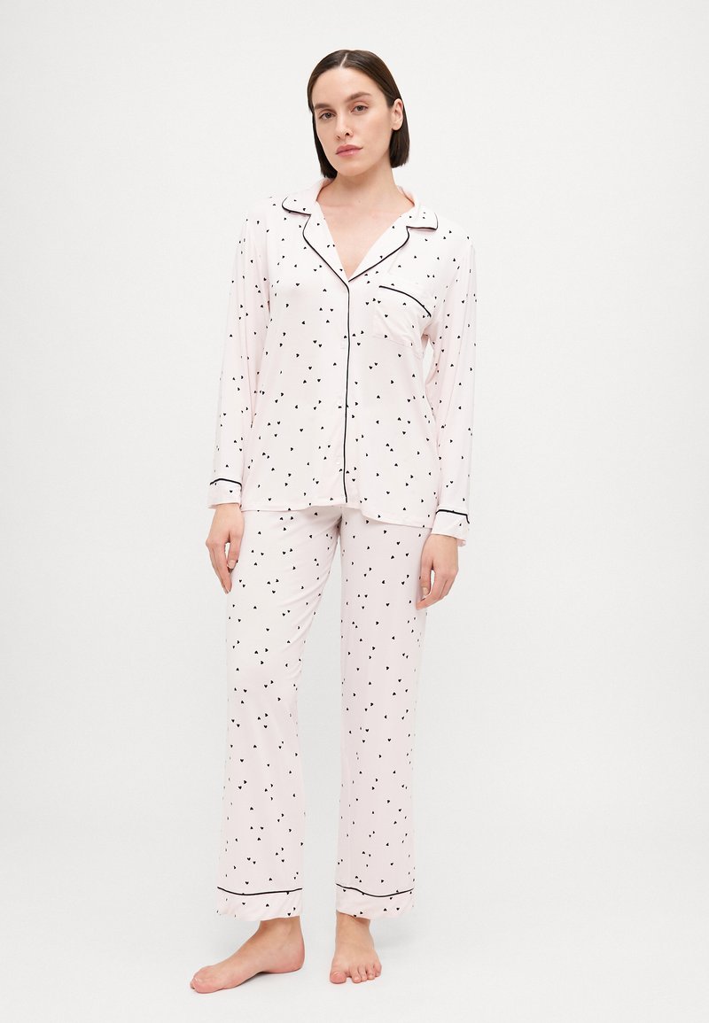 Ανοιχτό ροζ σετ πιτζάμας με μαύρα σχέδια καρδιών, μακριά μανίκια και μαύρες λεπτομέρειες. Το τοπ διαθέτει τσέπη; τα παντελόνια είναι φαρδιά.