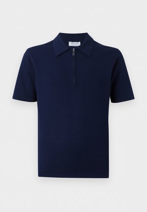 Homme aux cheveux bouclés portant une chemise bleu marine à manches courtes zippée, un pantalon beige à carreaux et des chaussures en cuir noires, debout les mains dans les poches.