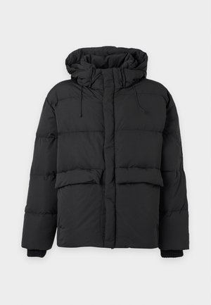 Ung mand iført en sort hættetrøje med dunjakke, hvid t-shirt, lyseblå løstsiddende jeans og røde sneakers, stående mod en ensfarvet baggrund.