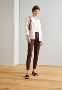 Chemise blanche à boutons, pantalon brun ajusté, sandales à talons noires avec un détail en nœud, et un sac à épaule marron. Fond de sol en bois.