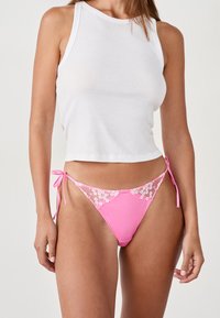 Bas de bikini rose avec des bordures en dentelle et des côtés à nouer, présentant des motifs floraux sur les bords. Associé à un haut sans manches blanc.