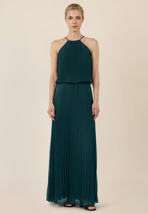 Kobieta w bezrękawowej, ciemnozielonej plisowanej sukience z dekoltem halter, stojąca na tle jednolitego beżowego tła.