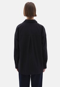 Zwarte katoenen shirt met een rugplooi, kraaglijn, lange mouwen met knoopmanchetten en een rechte onderkant.