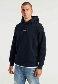 Felpa con cappuccio blu navy, dotata di tasca frontale, lacci e un piccolo logo. Tessuto morbido con polsini e orlo a costine. Indossata con jeans chiari.