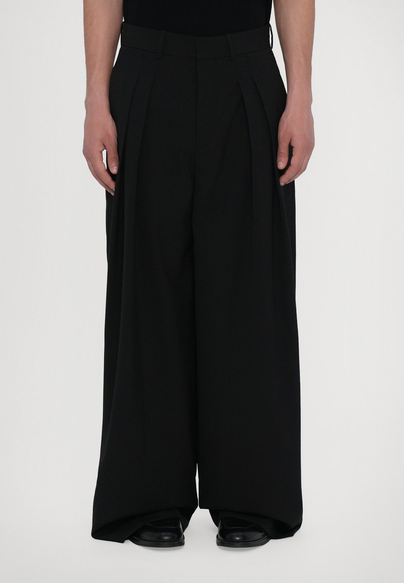 Black pleated trousers with a wide leg design, made from a smooth fabric. Features a fitted waist and is styled with black shoes.