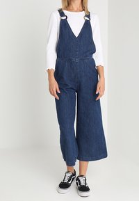 Denimöveralls med vida ben, justerbara axelremmar och sidofickor. Matchas med en vit långärmad topp och svarta sneakers.