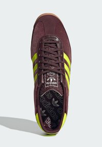 Burgundi bársonyos sportcipő három neon sárga csíkkal, texturált fűzővel és mintázott fekete talpbetéttel, amelyen az Adidas logo található.