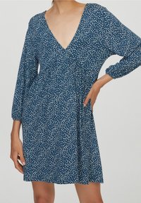 Robe fleurie bleue avec un décolleté en V profond, manches longues et coupe ample. Fabriquée en tissu léger avec un motif floral blanc.