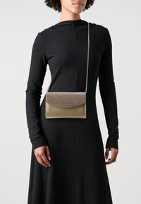 Złota kopertówka z teksturowanym, błyszczącym zapięciem, posiadająca gładkie złote body i łańcuszkowy pasek, noszona na czarnej sukience z długim rękawem.