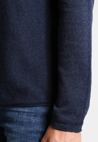 Maglione texture blu navy con polsini a coste, indossato su jeans in denim. Il tessuto appare morbido e leggermente elasticizzato.