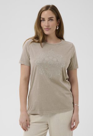 Donna con capelli castano chiaro lunghi che indossa una maglietta beige a maniche corte con un disegno mandala argentato borchiato e pantaloni chiari, in piedi contro uno sfondo bianco.