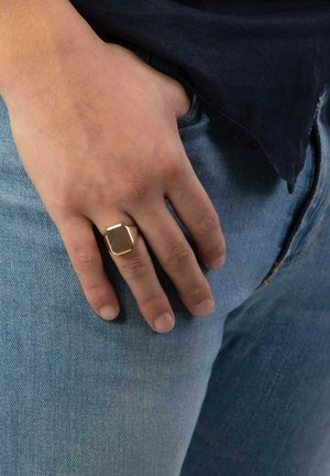 Gouden geometrische ring die gedragen wordt aan een hand die op denim rust. De ring heeft een hexagonaal ontwerp met een gestructureerde oppervlakte.