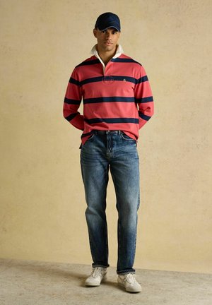 Muž oblečený v červeno-námorníckom pruhovanom ragby tričku, modrých džínsach, bielych teniskách a námorníckej šiltovke stojaci pred béžovým pozadím.