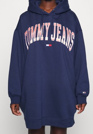 Person iført en oversized marineblå hættetrøje-kjole med tekst og logo "Tommy Jeans" på brystet og ærmet.