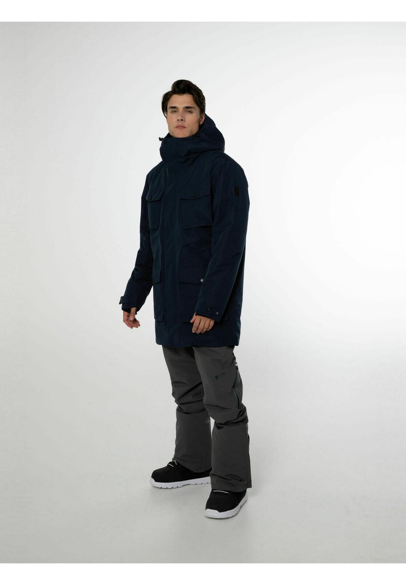 Parka bleu marine avec poches poitrine doubles, un col haut et une capuche, associé à un pantalon de neige gris et des chaussures noires.