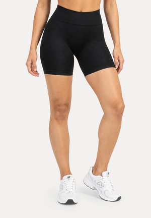 Femme portant un short cycliste noir taille haute et des chaussures de course blanches New Balance, debout devant un fond uni.