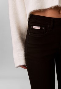 Melnas džinsa bikses ar pieguļošu dizainu, ar Calvin Klein logotipa tagu priekšējā kabatā. Džemperis ir balts un ar tekstūru.