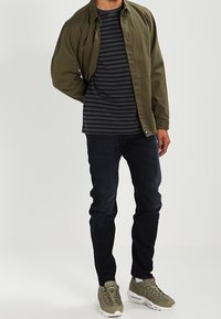 Chemise vert olive portée sur un t-shirt à rayures bleu marine et grises, associée à un jean foncé et des baskets olive avec des détails texturés.