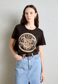 T-shirt nera con un design circolare di farfalla oro e rosa, abbinata a una gonna di jeans leggera blu e una cintura nera.