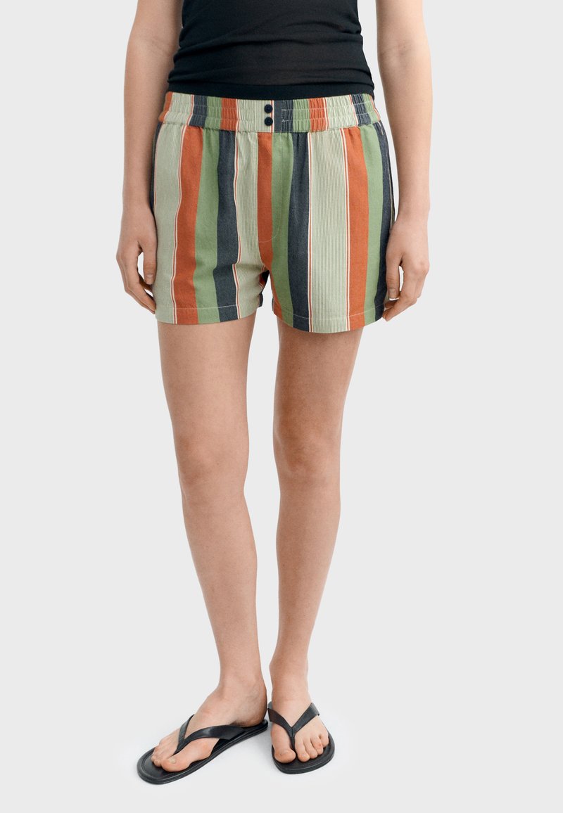 Personne portant un short ample à rayures verticales orange, vertes et noires, un débardeur noir et des tongs noires, debout devant un fond blanc.