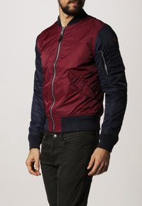 Burgundy och marinblå bomberjacka med dragkedja, sidofickor och ribbade muddar. Har en texturerad yta och en patch på ärmen.