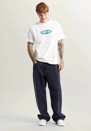 Hvid bomuld T-shirt med et blåt og grønt "vans" logo, kombineret med blå denim bukser og hvide sneakers. Har en afslappet pasform og korte ærmer.