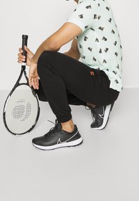 Muž v dřepu, oblečený v černých teniskách Nike a černých kalhotách, držící černobílou tenisovou raketu, má na sobě bílé tričko s potiskem psů.
