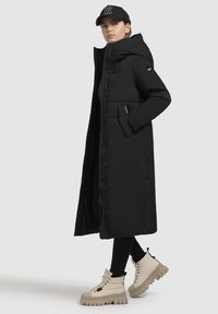 Crni dugi puffer kaput s kapuljačom, s prednjim gumbima, bočnim džepovima i elegantnim dizajnom. Uparen sa kremastim čizmama s visokim potplatom.