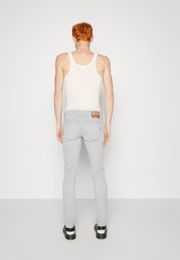 Hombre con cabello rojo que lleva una camiseta sin mangas blanca, jeans grises claros con bolsillos traseros, calcetines blancos y zapatos negros, de pie contra un fondo sencillo.