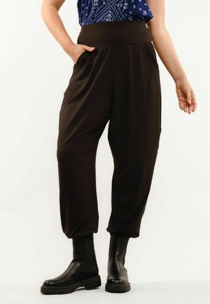 Osoba nosiaca čierne voľné nohavice s vysokým pásom a vreckami, čierne masívne čižmy a modrý vzorovaný top, stojaca na bielom pozadí.