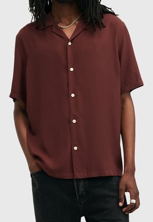 Man wearing a burgundy short-sleeve button-up shirt with cream buttons, a chain necklace, black pants, and a silver ring on his right hand.