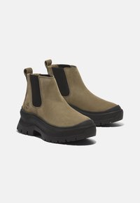 Olivgröna ankelstövlar med svarta elastiska sidoflikar och en kraftig svart gummisula. Slät mocka-textur, rund tådesign, logotypdetalj.