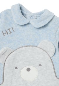 Monosie de peluche azul claro con un diseño de oso; presenta un "¡HOLA!" bordado y acentos en gris y blanco, con un cuello redondeado.