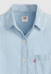Camicia di jeans blu chiaro con chiusura a bottoni, colletto a punta, tasca sul petto e il caratteristico gancio rosso Levi's. Presenta bottoni color avorio e una consistenza morbida.