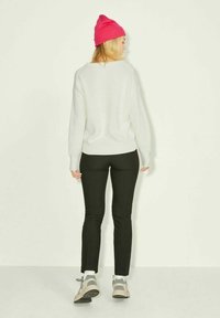 Femme de dos portant un pull en maille blanc, un pantalon noir, des baskets grises, des chaussettes blanches et un bonnet rose vif sur un fond uni.