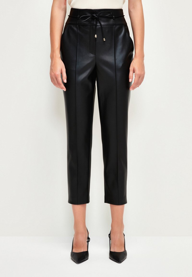 Femme portant un pantalon en cuir noir court avec des coutures avant et une ceinture nouée, associé à des talons hauts noirs pointus.